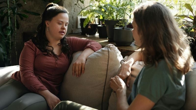 Two women sit together on a beige sofa in a sunlit living room with potted plants in the background, having a serious and engaged conversation. One woman wears a burgundy long-sleeved top whilst the other wears a green t-shirt, representing the important discussions parents and young people need to have about understanding the Renters' Rights Act and what changes mean for young people entering the private rental market.