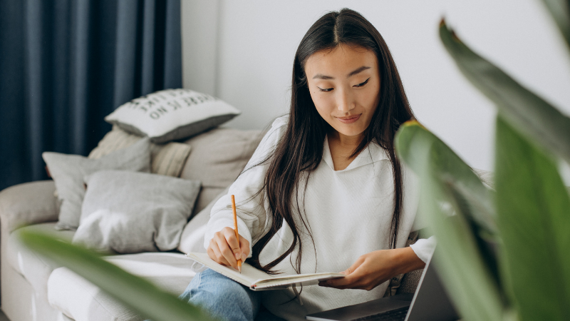 A young Asian woman with long dark hair sits on a bed with grey and cream pillows, wearing a white long-sleeved top and light blue jeans. She concentrates on writing in a notebook with a pencil, with a laptop beside her in her bright, minimally decorated bedroom featuring a blue curtain and green potted plants. The focused scene captures the practical preparation and planning involved when studying abroad, highlighting the important steps such as securing accommodation and understanding rental requirements in a new country.