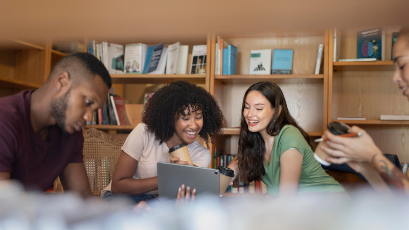 A diverse group of students gathered in a bright, modern library space with wooden bookshelves filled with books visible in the background. In the foreground, a woman with curly dark hair wearing a beige top and a woman with long dark hair in a green top smile as they look at a tablet together, engaged in collaborative learning. Other students are visible working in the background. The warm, inclusive scene captures the student experience and community aspect that parents should consider when understanding the student renting guide, illustrating how accommodation choices impact both academic success and social development during university.