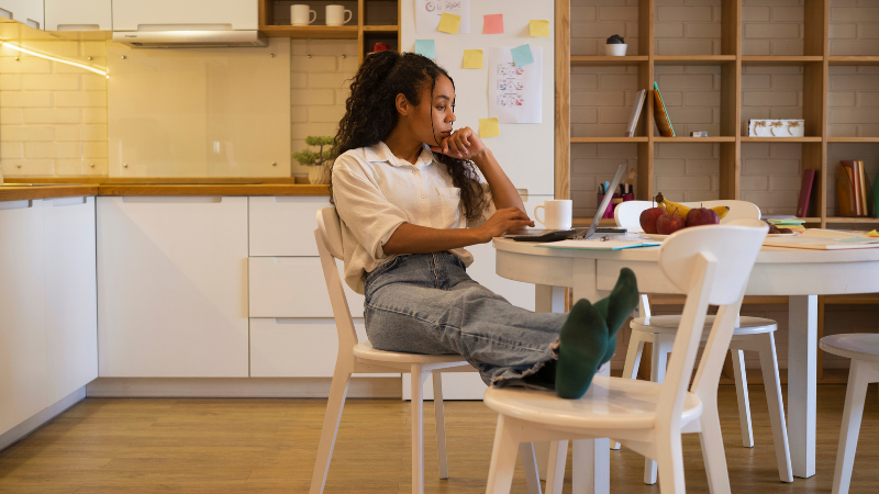 A young woman with curly dark hair, wearing a cream coloured shirt and grey jeans, sits thoughtfully at a modern white dining table in a bright, contemporary shared kitchen. She rests her hand against her face with a contemplative expression, surrounded by books, a cup, and an apple on the table. The minimalist kitchen features white cabinetry, wooden shelving with decorative items, and sticky notes on the wall. The scene captures the consideration and decision-making process students go through when comparing private rented accommodation versus purpose-built student accommodation, illustrating the moment of weighing up which option is right for them.