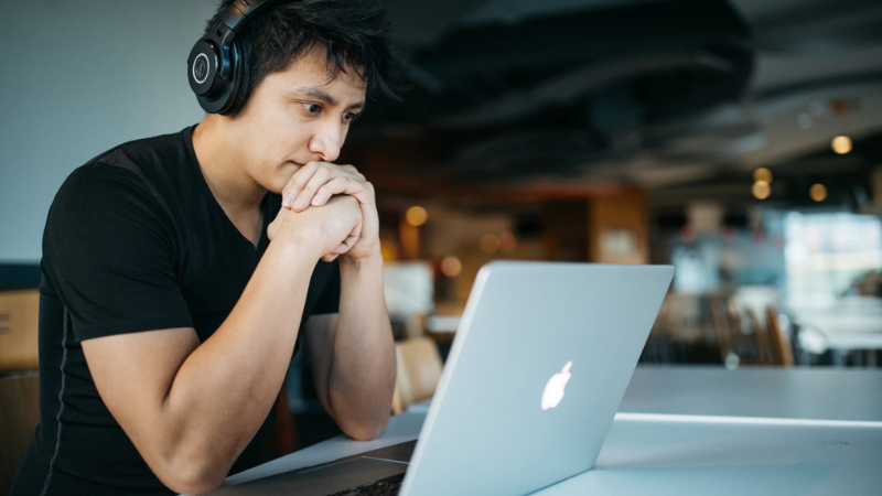 A young man wearing a black t-shirt and black over-ear headphones sits at a table with a silver laptop, his hand raised thoughtfully to his face as he considers the screen. The modern, brightly lit study space with soft bokeh lighting in the background captures the moment of research and contemplation students go through when navigating UCAS clearing, understanding what it is and how it works as they finalise their university choices.
