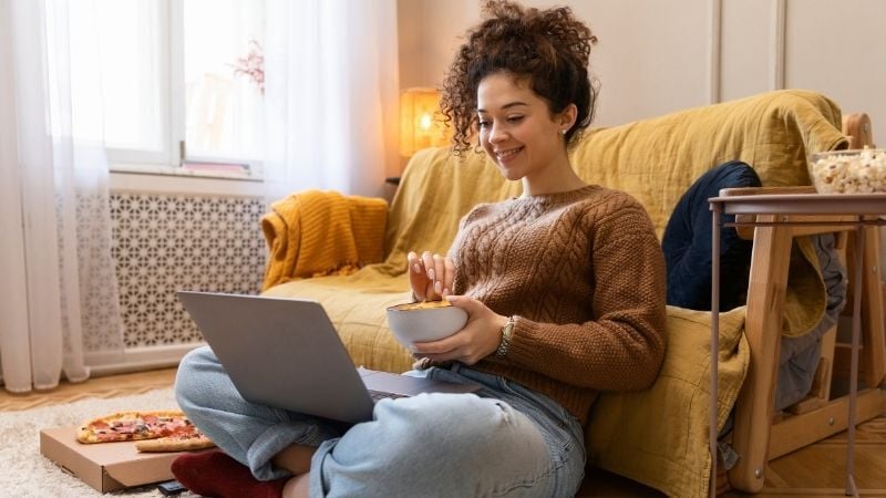 A young woman with curly dark hair sits on a mustard yellow sofa in a warm, cosy bedroom, wearing a brown cable knit sweater and light blue jeans. She holds a laptop whilst eating from a bowl, with a pizza box on the floor beside her, representing a student living independently. The comfortable, casual scene captures the reality of young people's student life and illustrates why parents need to understand what it means to be a guarantor for your young person when they move into private rented accommodation.