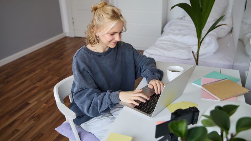 A young woman with blonde wavy hair, wearing a dark navy sweatshirt, sits at a white desk working on her laptop with a focused expression. The bright, modern workspace features colourful notepads in green, pink and yellow, a white mug, and a potted plant in the background, with wooden flooring visible. The scene represents someone researching and understanding what a guarantor passport is and how the pre-approved guarantor service works to streamline the rental application process.