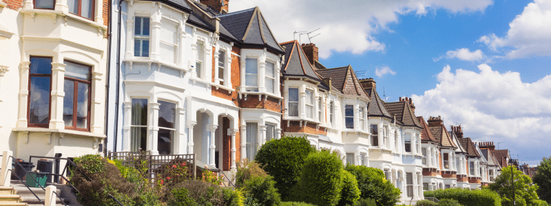 A row of Victorian terraced houses with white facades, bay windows, and brown tile roofs stretching into the distance. The houses feature characteristic period architecture with ornate details, small front gardens with green shrubs and trees, and black railings. The scene is set against a bright blue sky with white clouds, typical of British residential streetscapes.