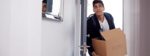 A young man with dark hair wearing a navy jacket over a white t-shirt is stepping through a white doorway while carrying a brown cardboard moving box labeled "KITCHEN." He appears to be looking upward as he navigates through the entrance. The scene takes place in a bright, minimally decorated room with white walls and a mirror visible on the wall, capturing the moment of moving into new accommodation and illustrating the practical steps involved in following a student moving checklist.
