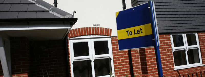 13:51A blue and yellow "To Let" sign stands prominently in front of a modern brick-built residential property with white-framed windows and a tiled roof under an overcast sky. The letting board represents the competitive UK rental market where the demand for guarantors continues to grow as landlords seek financial security when letting properties to tenants without traditional proof of income or UK-based family support.