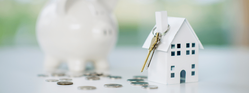 A photograph showing a white miniature house model with a key attached, positioned next to a blurred piggy bank and scattered coins on a light surface with a soft, bright background. This image represents the various fees when renting in the UK, including deposits, guarantor fees, and other rental costs that tenants need to budget for when securing accommodation.