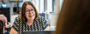 A woman with shoulder-length brown hair and glasses, wearing a black and white geometric patterned top, sits at a table with a laptop and coffee cup in a modern office or café setting. She has a warm, engaged expression as she appears to be in conversation, representing someone considering the responsibilities and requirements of who can be a rent guarantor for a family member or friend seeking rental accommodation.