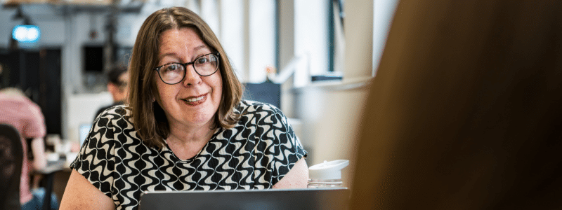 A woman with shoulder-length brown hair and glasses, wearing a black and white geometric patterned top, sits at a table with a laptop and coffee cup in a modern office or café setting. She has a warm, engaged expression as she appears to be in conversation, representing someone considering the responsibilities and requirements of who can be a rent guarantor for a family member or friend seeking rental accommodation.