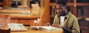 A focused young Black man in an olive green jacket over a white t-shirt sits at a wooden table in a traditional library, intently reading an open book with stacked books and notebooks beside him. The warm, academic setting with wooden bookshelves in the background captures the atmosphere and educational opportunities of studying in London, including places like the British Library.