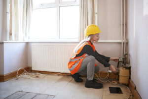 A photograph of a female construction worker or tradesperson wearing a yellow hard hat and orange high-visibility vest, crouched down working on plumbing or heating installations near a radiator in an unfurnished room. The person is positioned by a window with white curtains, and there are tools and equipment scattered on the wooden floor,