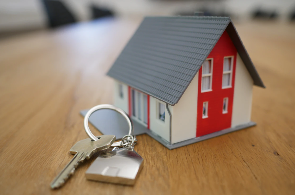 A close-up photograph of a miniature house model with red and white walls and a grey roof, positioned next to a set of keys with a house-shaped keyring on a wooden surface.