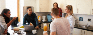 A group of five students gathered in a bright, modern shared kitchen with white cabinets, having a discussion around a laptop on the dining table. The diverse group includes students working together, with drinks and notebooks visible, representing housemates collaborating and making important decisions about finding suitable second year accommodation after their first year in university halls.