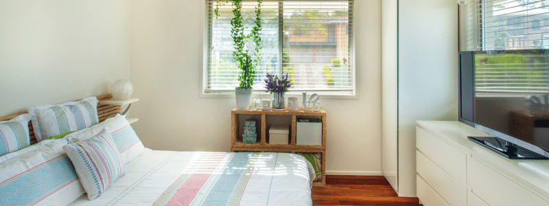A bright, well-decorated student bedroom featuring colorful striped bedding, natural light from large windows, wooden furniture, and potted plants, demonstrating creative and affordable ways to decorate your student room on a budget while creating a comfortable living space.