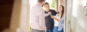 A professional letting agent or landlord in a pink shirt, viewed from behind, hands over keys to a smiling young couple at the doorway of a bright rental property. The happy tenants - a man in a dark t-shirt and a woman in a blue and white striped shirt - receive the keys, representing the successful letting process where landlords have considered financial protection options like rent guarantee insurance vs rent guarantor to safeguard their rental income.