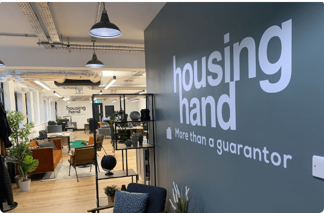Interior view of the Housing Hand office showing a modern open-plan workspace with exposed ceiling elements, industrial pendant lighting, and wooden floors. A prominent dark grey feature wall displays the company name 'housing hand' in large white lowercase letters with the tagline 'More than a guarantor' below. The space includes contemporary furniture, plants, shelving units, and workstations, creating a welcoming professional environment.
