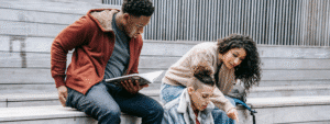 Three diverse students sitting together on outdoor steps, with one reading from a book while the others listen and engage in discussion, representing the collaborative learning experience and community aspect of choosing to study in the UK.