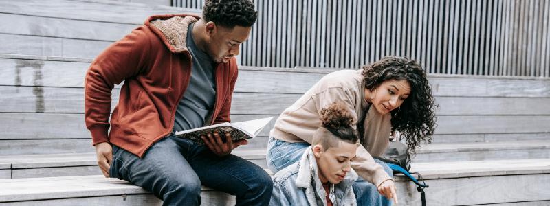 Three diverse students sitting together on outdoor steps, with one reading from a book while the others listen and engage in discussion, representing the collaborative learning experience and community aspect of choosing to study in the UK.