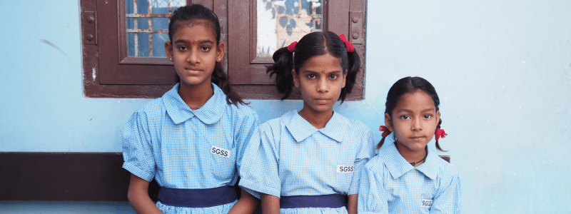 17:04Three young Indian girls in matching light blue gingham school uniforms with 'SOSS' badges stand together outside a building with weathered blue and brown windows. The girls, of varying ages with their hair styled in plaits and ponytails adorned with red ribbons, look directly at the camera representing the educational opportunities and life-changing impact that charitable initiatives can provide to transform lives of three Indian girls and support their futures.