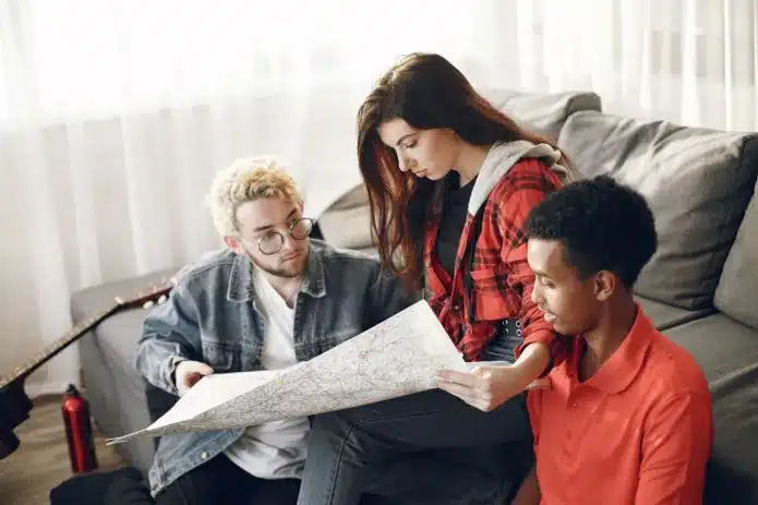 A photograph of three young people - a perso with short blonde hair in a denim jacket, a person with brunette hair in a red and black checked shirt, and a person in an orange jumper - sitting together on a sofa examining documents or paperwork. The scene appears to be in a bright, modern living space with large windows, suggesting they may be reviewing rental agreements, housing contracts, or property-related documents together