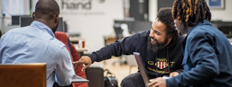 A photograph showing three people in what appears to be an office or meeting space, with a man in the centre wearing a dark hoodie, engaged in conversation with two other individuals - one in a light blue shirt and another in dark clothing. The scene suggests a consultation or advisory meeting, relating to how to find a guarantor services where people seek guidance on securing rental guarantors for housing applications