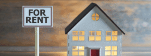 A miniature grey house model with a dark roof and illuminated yellow windows, positioned next to a white 'FOR RENT' sign on a post. The scene is photographed with a soft, blurred background suggesting a rental property market context. This image relates to rental guarantor services, explaining what is a guarantor for rent - typically someone who agrees to cover rental payments if a tenant cannot meet their obligations.