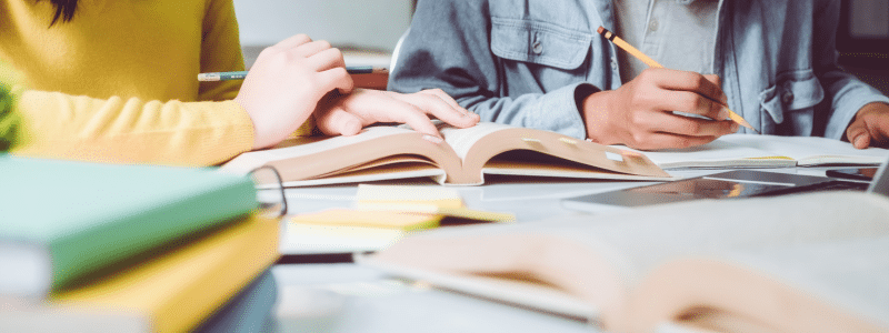 A close-up photograph of two people studying or working together at a table, with one person wearing a yellow jumper and another in a light blue shirt holding a pencil. The scene shows open books, notebooks, and a stack of coloured folders or binders in the foreground, creating a warm, educational atmosphere with soft natural lighting.