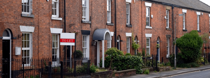 A row of traditional red brick Victorian terraced houses with white window frames and black iron railings, featuring a prominent "TO LET" sign in the foreground, illustrating the typical rental property market and housing options available when renting in the UK