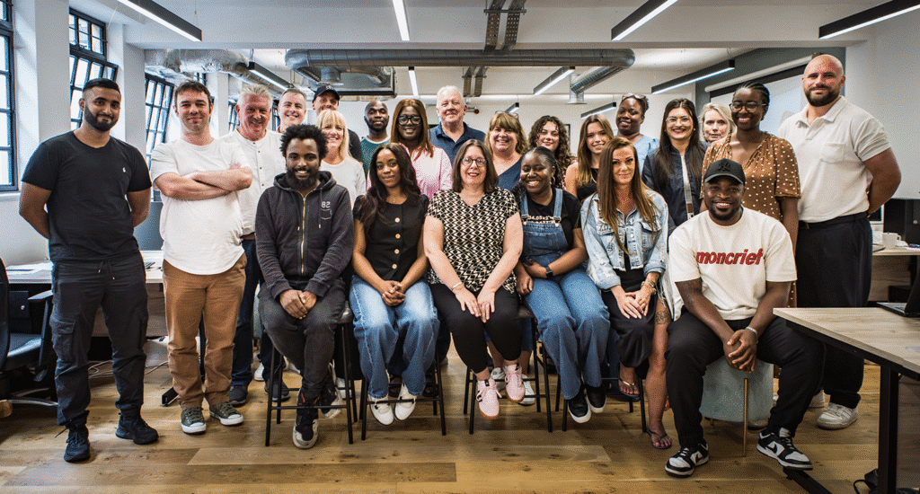 A group photograph of the HousingHand team. There are over 20 members of their diverse team members in a modern office space with exposed ceiling beams, large windows, and wooden floors. The team includes men and women of various ages and ethnicities, dressed in casual business attire, arranged in two rows with some seated and others standing.