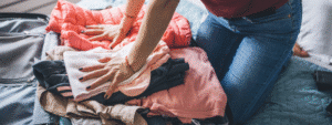 A photograph showing someone wearing jeans kneeling on a bed whilst folding and organising colourful clothing including coral, white, and dark-coloured garments. The scene depicts the process of packing for university, with various items of clothing being sorted and prepared for student accommodation