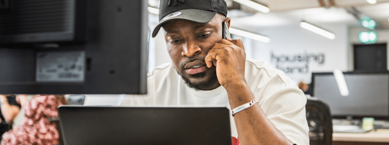 A photograph of a man wearing a grey baseball cap and white shirt, speaking on a mobile phone while looking at a laptop screen in what appears to be a modern office environment with blurred signage for Housing Hand in the background. This image relates to professional guarantor services, where individuals can access expert support and guidance when securing rental housing in the UK.
