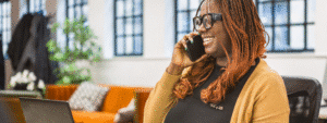 A smiling woman with glasses and dreadlocks talking on her phone in a bright, modern office space with large windows, representing accessible healthcare communication and the type of comprehensive support for tenants that services like Housing Hand's 24/7 GP and wellbeing programs provide.