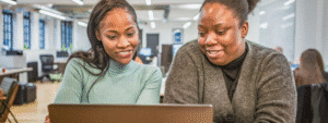 Two smiling women looking at a laptop together in a modern office environment, representing students researching and collaborating on important decisions such as how to choose student accommodation that best fits their needs and budget.