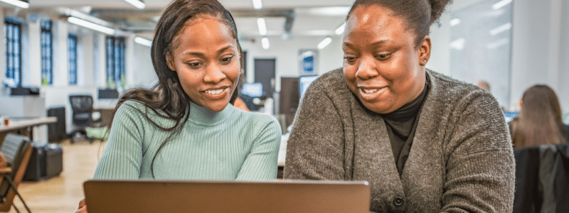 Two smiling women looking at a laptop together in a modern office environment, representing students researching and collaborating on important decisions such as how to choose student accommodation that best fits their needs and budget.