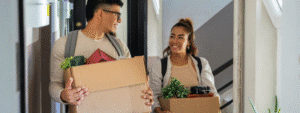 A young man wearing glasses and a young woman, both carrying cardboard boxes with small potted plants and personal belongings, smile at each other as they move into a shared property. The scene in a modern hallway captures the optimistic start of shared tenancy, highlighting the importance of understanding joint and several liability in rental contracts to avoid being financially responsible for flatmates' unpaid rent.