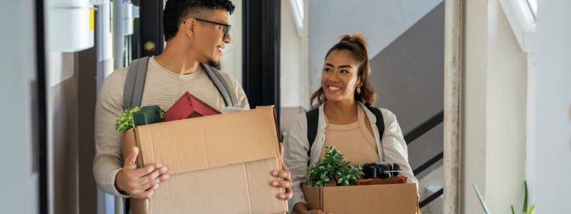 A young man wearing glasses and a young woman, both carrying cardboard boxes with small potted plants and personal belongings, smile at each other as they move into a shared property. The scene in a modern hallway captures the optimistic start of shared tenancy, highlighting the importance of understanding joint and several liability in rental contracts to avoid being financially responsible for flatmates' unpaid rent.