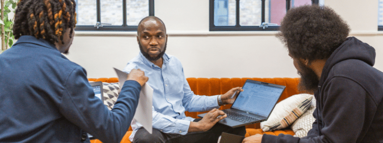 Three people sitting on an orange upholstered bench seat in a bright, modern office space with large windows. A professional in a light blue shirt sits in the centre holding a laptop, engaged in discussion with two others - one in a dark jacket gesturing with their hand and another in a black hoodie. The collaborative meeting scene represents landlords, letting agents, or tenants discussing the implications and changes introduced by the Renters' Rights Act and how it affects rental agreements and tenant protections.